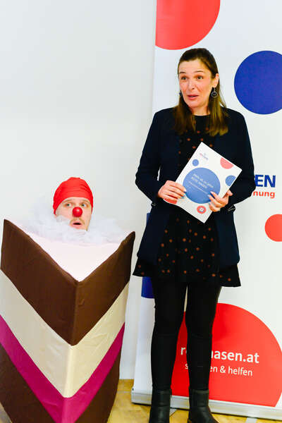 Ein Clown, versteckt in einem Tortenstück und Frau Janine Vasiri, die eine Rede hält.