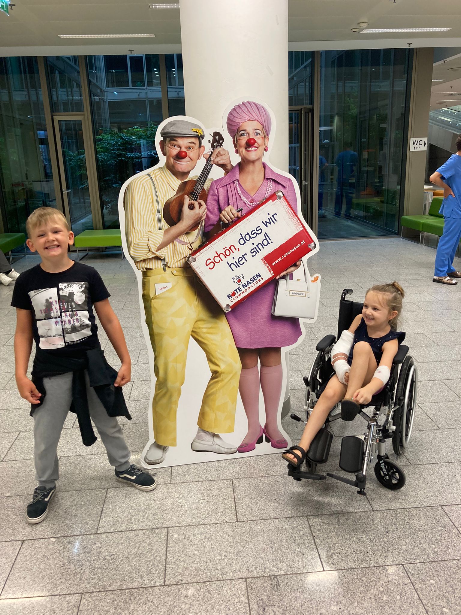 Frida und ihr Bruder im Spital vor einem Pappaufsteller mit zwei ROTE NASEN Clowns.